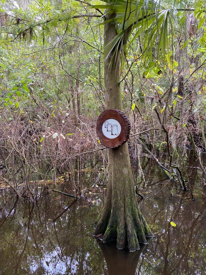 Wood-burned Stump Jumper Pass marker sign — a round wood slice with an etched stump illustration, mounted on a cypress tree rising from flood water